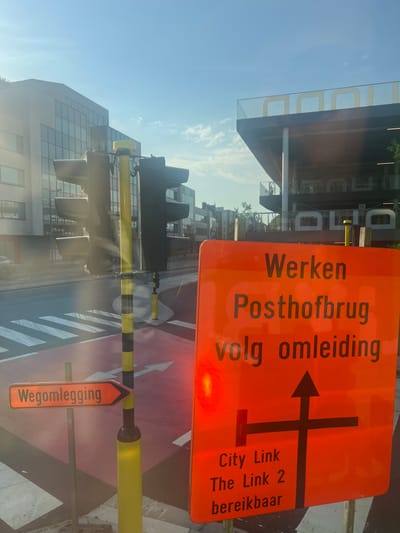 The image depicts a street scene in Antwerp, Belgium, dominated by an orange road construction sign. The sign reads "Werken Posthofbrug volg omleiding," meaning "Work Posthof Bridge follow detour." Below this, a diagram indicates a detour route with arrows showing a straight path, a right turn, and a left turn. Further text on the sign states "City Link The Link 2 bereikbaar," indicating that City Link and The Link 2 are accessible.
In the foreground, a yellow traffic signal pole with black bands stands prominently, partially obscured by sunlight glare. A smaller orange directional sign with the text "Wegomlegging" (detour) points to the right. The ground features a red-marked pedestrian crossing and a bike lane.
In the background, modern buildings with glass facades and a structure with large yellow lettering are visible under a clear blue sky with scattered clouds. The overall scene suggests active road construction and traffic management in an urban setting. There are no people or vehicles clearly visible in the image.
