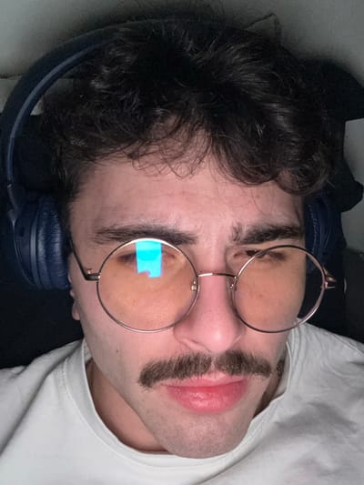 A man lies on his side, resting his head on blue headphones. He has dark, curly hair and wears round, wire-rimmed glasses with a light reflection on one lens. A thick, neatly trimmed mustache sits above full lips. He wears a white t-shirt. The background includes dark fabric and possibly a pillow, suggesting he's lying down to relax.