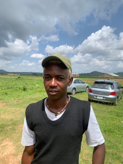 A young man with blonde-tipped hair and a green cap stands in a grassy field with two cars parked behind him. He is wearing a white t-shirt and a black sleeveless sweater, and a beaded necklace with a cross pendant. The sky is cloudy with patches of blue. The location is identified as Gwafan, Nigeria.