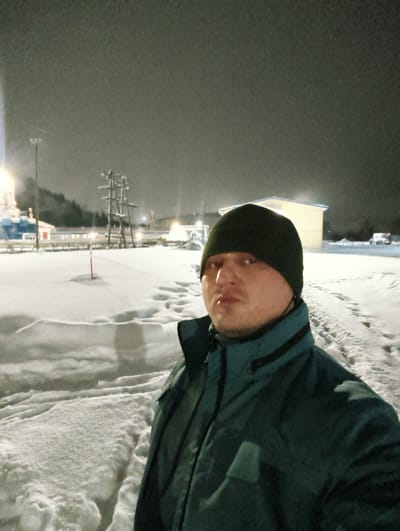 The image captures a male individual at night in the city of Im. Gastello, Russia. The man occupies the foreground, facing the camera directly, wearing a dark green beanie and a dark teal winter jacket with a black collar and black trim. The ground is extensively covered in snow, exhibiting visible footprints and disturbed areas. In the mid-ground, a tall, snow-laden wooden structure or scaffolding is present. To the left, an industrial area is illuminated by bright lights, revealing a large vessel, possibly a ship, and utility poles. A light-colored building with a pale roof and white walls is situated in the middle-right background. Additional distant structures and snow-covered terrain are discernible against the dark night sky. Multiple artificial light sources cast illumination across the snowy environment.