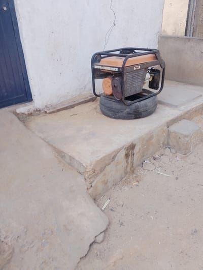 A Firman portable generator, featuring an orange fuel tank and a black metal frame, rests on a black rubber tire. The tire is positioned on a raised, multi-level concrete platform. The generator's engine block is grey, with an orange fan housing visible on its left side. Dark, vertical stains are present on the side of the concrete platform below the generator. Behind the platform, a textured white wall stands, partially revealing a dark blue door on its left side. A thin, dark wire or cable hangs vertically along the white wall. The ground in the foreground and to the right of the platform consists of dry, light-brown sandy soil, scattered with small pieces of light-colored debris. No individuals, actions, or interactions are present in this static scene, located in Dorayi, Nigeria.