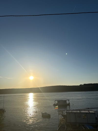 The image depicts a tranquil scene on the Danube River in Silistra, Bulgaria, during what appears to be sunset. The sun is low on the horizon, casting a warm, golden glow across the water and creating a shimmering reflection. A distant, tree-lined shore forms a dark silhouette against the bright sky.
In the foreground, several structures are moored on the river. One is a houseboat with a Bulgarian flag flying from its mast, indicating a connection to the country's identity. Next to it, another floating structure, possibly a small cabin or utility building, is also visible. Several small boats are scattered in the water, some tied to the structures and others adrift.
The sky is a clear gradient of blue and yellow, with the sun being the dominant light source. A thin, dark power line stretches horizontally across the upper portion of the frame, a man-made element contrasting with the natural beauty of the river and sky. The overall impression is one of peacefulness and stillness, characteristic of a quiet evening by the water.