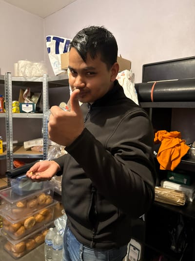 A male individual is depicted indoors within a storage or utility room in Holtzheim, France. The man, with short dark hair, wears a black zip-up jacket and blue jeans. He faces forward with a slight smile and narrowed eyes, displaying a "thumbs up" gesture with his right hand. His left hand is cupped, holding a small quantity of mixed nuts or seeds.

The background features a silver metal shelving unit on the left, stocked with various items including rolls of white material, a white plastic bag with blue text, cardboard boxes, packaged goods, and stacked clear plastic containers filled with bread rolls. Two clear plastic water bottles rest on the floor below. On the right, a black shelving unit holds a rolled black mat secured with an orange strap, a black case, an orange folded garment, a small green box, and bundles of thin packaged sticks. The room walls are light-colored.