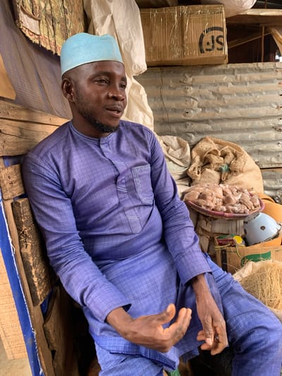 This media file shows a medium shot of a Black man wearing a blue skullcap and a purple patterned tunic, lying on his back. He appears to be in an outdoor or semi-outdoor setting, possibly a market or storage area, given the corrugated metal walls and stacks of goods in the background. To his left, there are cardboard boxes with the "SF" logo visible, wrapped in plastic. To his right, various items are stacked, including a woven basket filled with small, individually wrapped items, a large sack, and some straw-like material. The lighting suggests it could be daytime. The man's gaze is directed upwards and slightly to his left, and his expression appears neutral. His hands are resting on his chest and abdomen.