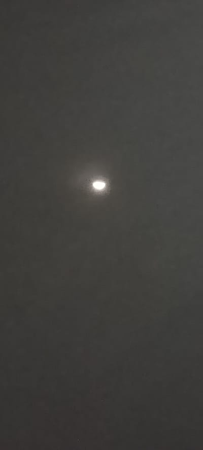 The image is a dimly lit, vertical photograph of a dark, textured surface, possibly a wall or fabric, with a blurred, bright, semi-circular light source positioned slightly above and to the left of the center. The light source appears to be a celestial body, likely the moon partially obscured by clouds or haze, or possibly a strong artificial light. There are no people, specific objects, or discernible environmental features present in the image to indicate its location in Sihomlhok, Indonesia. The overall impression is one of muted tones and a sense of distance or obstruction of the light.