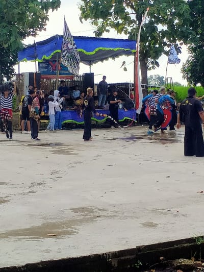The image shows a performance taking place in Kecamatan Kedawung, Indonesia, at coordinates -7.519, 111.028. A group of people are gathered in front of a stage decorated with blue and white fabric and flags. The stage has a large banner with the word "LEGENDARY" printed on it.  Some individuals are standing on the stage, while others are in front of it. There is a man in a black outfit holding a long, flexible rod. He is positioned in front of the stage, and is the focal point of the scene.  Several other individuals are dancing in front of the stage. They are dressed in various colors, including black, white, and red. Some are holding flags and other objects, while others are simply moving their bodies to the music.  The environment is outdoor, with a concrete surface and some trees in the background. It seems that it may have rained recently as there are puddles on the ground and it is possible that the event is taking place after a downpour.  The overall atmosphere of the image is festive and celebratory.