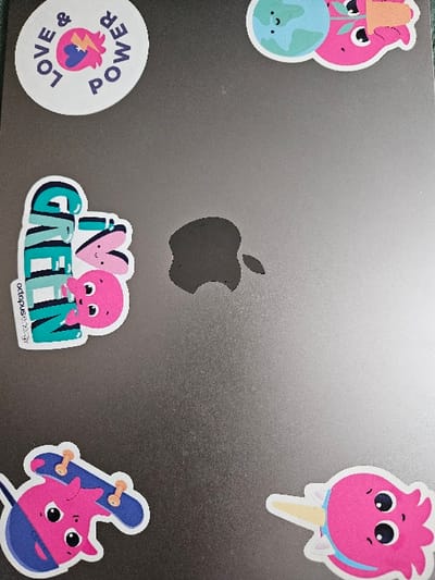 The scene displays the gray lid of an Apple MacBook laptop, identifiable by its central black Apple logo. The surface is adorned with five distinct stickers. In the upper left quadrant, a circular white sticker reads "LOVE & POWER" in blue text, surrounding a stylized pink heart with a blue lightning bolt. To the upper right, a sticker depicts a pink octopus-like character holding a small green plant in an orange pot, positioned beside a light blue globe with a smiling face. On the middle left, a sticker features a pink octopus-like character with white bunny ears partially obscured by the teal word "GREEN," with "octopus.energy" visible beneath. The lower left area holds a sticker of a pink octopus-like character wearing a blue cap and riding a blue skateboard. In the lower right, a sticker shows a pink octopus-like character with a blue stripe and a light yellow cone, resembling an ice cream. The overall environment is indistinct, suggesting an indoor setting within the Cherwell District, United Kingdom.