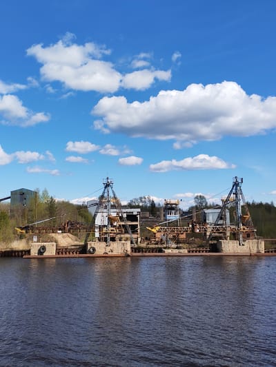 The image depicts an industrial waterfront scene in Ivanov Bor, Russia. Two large, decaying crane structures with rusted metal frames and concrete bases are situated on what appears to be a dilapidated dock or platform extending into a body of dark, rippling water. Old tires are fastened to the dock's edge. Behind the cranes, amidst vegetation and trees, are other industrial buildings, including a structure with a conveyor belt. The sky is bright blue with puffy white clouds. The overall impression is one of disuse, obsolescence, and industrial decline.