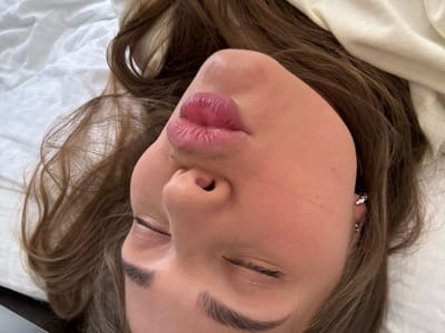 A person with fair skin and light brown hair is lying on a white sheet. Their eyes are closed, and their lips are pursed. The person is wearing earrings in their left ear. The background is blurry and appears to be a plain white surface. The location is not evident from the image. The image has no indications of being in Sosnovy, Russia.
