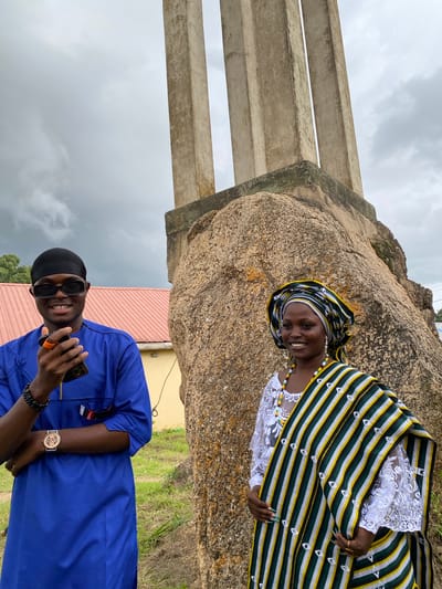The media file features two individuals in traditional Nigerian attire, posing in an outdoor setting. The scene is dominated by a large rock formation, with several concrete pillars visible to the left, suggesting a historical or monumental site.
The woman, positioned slightly behind and to the right, wears a vibrant striped headscarf and a white lace top. She is adorned with a colorful beaded necklace. Her expression is a joyful smile, and she is looking towards the camera.
The man, in the foreground, is dressed in a royal blue garment. He is wearing dark sunglasses and a black head wrap. He is holding a smartphone in his right hand and has a wrist watch and bracelet on his left arm. He is also smiling and looking at the camera, with a relaxed and happy demeanor.
The background includes grassy ground and a building with a red roof. The sky is overcast and appears to be a cloudy day. The lighting suggests it is daytime, possibly late morning or afternoon. The overall atmosphere is positive and celebratory, with the subjects appearing to enjoy their time together at this location in Wang, Nigeria. There is no visible text in the media file.