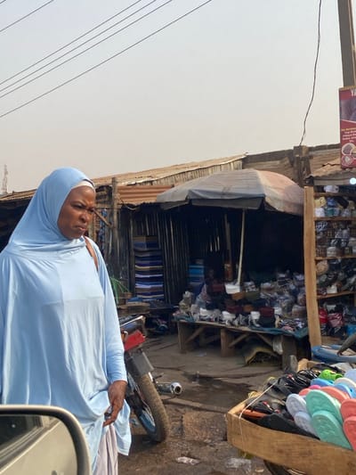 A woman in a light blue hijab and garment stands near a roadside shop. The shop displays stacks of various items, including footwear and fabric rolls, under a makeshift corrugated metal and tarp awning. A motorcycle is parked nearby. Power lines stretch across the sky. The scene appears to be in a bustling outdoor market area.
