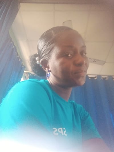 The image shows a woman with dark skin and black hair pulled back in a bun, wearing blue earrings and a blue t-shirt. The location is likely indoors, given the presence of a ceiling with a ceiling fan. Blue curtains are visible in the background. The environment appears to be illuminated by a light source in front. The presence of electrical wiring suggests the location is in Unguwar Rimi, Nigeria.