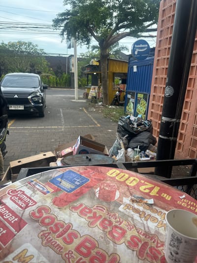 The image depicts a street scene in Celuk, Indonesia. A black car is parked along the side of the street with license plate "DK 1048 ACZ." In the background are businesses, including a Foodboxs restaurant, recognizable by its blue sign. Garbage is piled up on the side of the road, near some cardboard boxes. In the foreground is an outdoor table with a promotional sign for a food vendor, with logos for Indomaret and "Yummy choice," advertising "Sosis Bakar Ayam & Keju Melted Cheese" for Rp 12,000. A used paper cup sits on the table.
