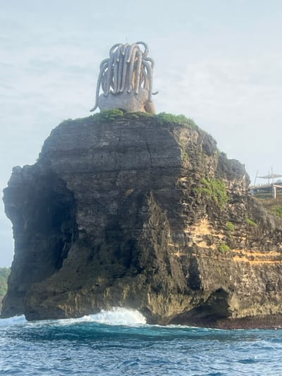 A large, weathered, dark gray rock formation rises steeply from the ocean. Perched atop this cliff is a light-colored, abstract sculpture resembling a stylized head with multiple thick, coiled or dreadlock-like appendages cascading down its sides. Green vegetation is visible on the upper surface of the cliff surrounding the sculpture. The cliff face itself exhibits horizontal stratification and varies in texture, with darker, smoother sections and rugged, eroded areas. The base of the cliff shows signs of significant erosion and undercutting by the sea, with a prominent wave crashing and producing white foam against the rock in the lower left quadrant of the image. The ocean water is a deep blue-green, exhibiting ripples and swells. In the upper right background, on a higher, vegetated landmass, a light-colored, open-air structure, possibly a bamboo or wooden platform, is partially visible. The sky above is a pale blue with minimal cloud cover. This scene is located in the city of Sakti, Indonesia. No individuals are present in the frame.