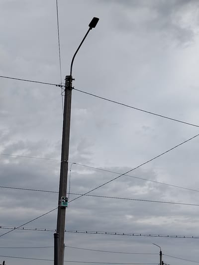 The image shows a concrete utility pole with a streetlamp attached to its curved arm, against a cloudy grey sky. Several dark electrical wires crisscross the frame, some attached to the pole. A small, white rectangular box with a green label and black text is mounted on the pole, approximately one-third of the way down from the top. The label on the box appears to have a drawing of an animal and the number "13344". The scene is likely an outdoor urban or suburban environment in Ekibastuz, Kazakhstan, indicated by the typical construction of utility poles and the presence of electrical infrastructure. There are no people visible in the image.