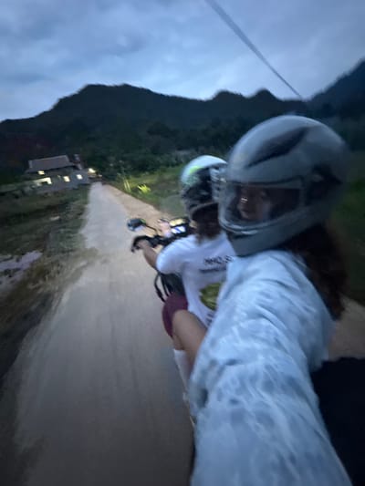 The image captures a low-angle, slightly blurred view from the perspective of someone riding on the back of a motorcycle, likely at dusk or early evening. Two individuals are visible, both wearing helmets. The person in the foreground, with a gray helmet, is wearing a light blue, possibly tie-dyed, t-shirt and has dark hair. Their right arm is visible, holding onto something, and their face is turned slightly away from the camera. Behind them, another person in a white t-shirt with a graphic on it and a lighter-colored helmet is seated in front.
They are traveling on a dirt or unpaved road that stretches into the distance. On the left side of the road, there's a traditional Vietnamese house with illuminated windows. Lush vegetation and dark, undulating hills or mountains form the backdrop. The sky is overcast and a deep blue-gray, suggesting twilight or cloudy conditions. The overall impression is of a rural setting in Mai Chau, Vietnam, with the journey taking place along a quiet, scenic route.