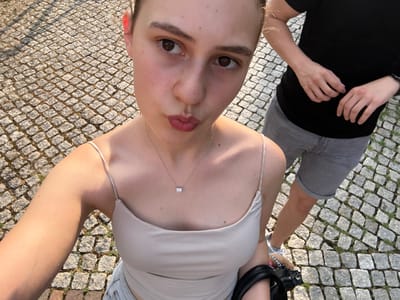 This media file is a selfie taken from a high-angle perspective, showing two people in an outdoor setting. The primary subject is a young woman in the foreground, with her face turned towards the camera. She is wearing a light-colored spaghetti strap top and a delicate necklace with a teardrop-shaped pendant. Her expression is neutral, with her lips slightly parted.
To the left of the woman, a second person, likely male, is partially visible. He is wearing grey shorts and a black t-shirt. His hands are resting on his lap, and he appears to be looking down.
The setting is a cobblestone ground, suggesting an urban or historical area. The lighting indicates it is daytime, with sunlight casting shadows on the ground. The overall impression is a casual moment shared between two individuals outdoors. The photo appears to be taken in Gernsbach, Germany, though there are no explicit text cues or landmarks to confirm this within the image itself.