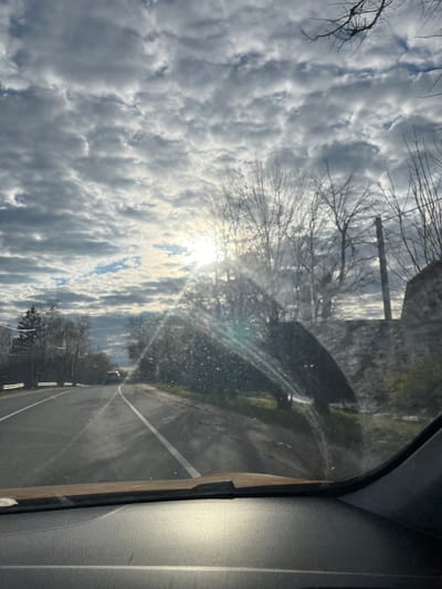The image, captured in Maksimkovo, Russia, is taken from inside a vehicle looking out through the windshield. The original photograph is rotated 90 degrees clockwise. When viewed with the sky at the top and the road at the bottom, the scene unfolds during daytime, likely morning or late afternoon, under partly cloudy conditions.
The upper portion of the frame is dominated by a textured, partly cloudy sky, featuring dense, grey and white stratocumulus or altocumulus clouds interspersed with brighter patches of clear sky. A prominent sunburst emanates from the upper-left, partially obscured by clouds, creating significant glare and lens flares across the upper-middle section of the image, indicating the sun's intensity.
Silhouetted against the bright sky are numerous bare branches of deciduous trees, suggesting a winter or early spring setting. In the mid-ground, along the sides of the road, more densely vegetated areas and possibly distant structures are visible, though somewhat indistinct due to the lighting and distance.
A paved road occupies the lower-middle portion of the image, stretching into the distance. It is marked with white lane lines, possibly a double solid line or a single line along its right edge, and its surface appears somewhat reflective.
On the far right, elements of the car's interior are visible, including a dark A-pillar and a portion of the textured dashboard or door panel, confirming the perspective from within a car. The windshield through which the photo was taken shows numerous small specks and streaks, likely dirt or water droplets, which contribute to the diffused light and scattered appearance of the glare.
The image captures a moment of travel, as if driving, with no discernible people or specific event taking place. There is no visible text in the image.