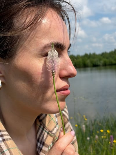 A young woman is pictured in profile, looking to the right, holding a small plant with a fuzzy, pale purple flower near her face. She has dark hair and is wearing a plaid shirt in shades of tan and brown. Her skin is lightly tanned, and the sunlight casts shadows across her face. In the background, a body of water stretches out, bordered by a line of green trees under a partly cloudy blue sky. The foreground also shows green grass and a scattering of small yellow and purple wildflowers. The scene appears to be a natural outdoor setting, possibly a park or lakeside area, with no urban elements visible. The image was taken in the city of Gushchino, Russia.
