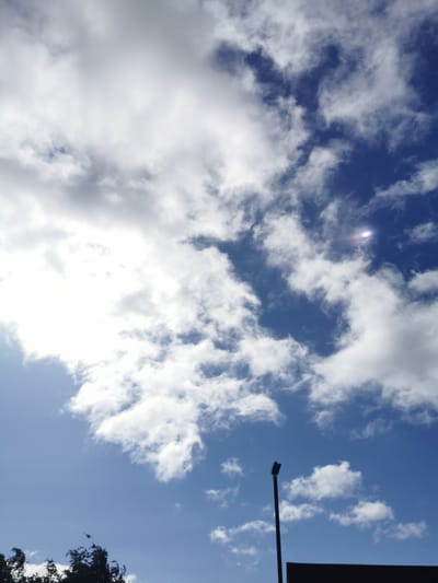 This is a vertical, eye-level, full shot of a bright blue sky filled with fluffy white and grey clouds. The sun is partially visible, creating a lens flare and a bright halo effect. In the lower third of the image, a tall, dark, thin lamppost with a light fixture at the top stands on the right, silhouetted against the sky. Below the lamppost, the dark silhouette of a building's roof is visible. In the bottom left corner, the dark, silhouetted branches of trees are visible. The overall impression is of a cloudy but bright day. The weather appears to be partly cloudy. The scene suggests an outdoor setting, possibly urban or suburban, given the presence of the lamppost and building. The time of day appears to be daytime, likely late morning or early afternoon, judging by the brightness and the sun's position. There is no visible text in the image.