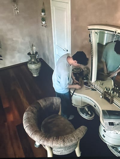 The image depicts an indoor scene, possibly a dressing room or bedroom, with a person actively engaged with items on a vanity. The room features a large mirror on the vanity, reflecting the person. In the foreground, a plush, oversized armchair is visible. The walls have a textured finish. A large vase stands on a dark wooden surface to the left of the frame. The overall impression is that of a private, domestic space. The location in Dondușeni, Moldova, cannot be determined from the visual information provided in the image.