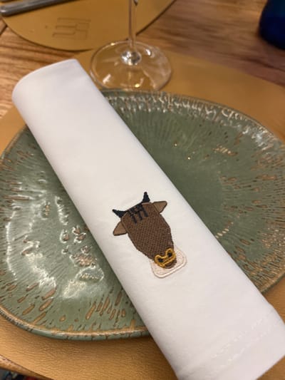 A white linen napkin with a brown bull embroidered on it is laying on a green ceramic plate, which is resting on a brown wooden table. There is a stemless wine glass, upside down, on the table to the left of the napkin. The coordinates 28.102, -15.414 correspond to the city of Las Palmas de Gran Canaria, Spain. The image is taken indoors, likely at a restaurant or café.