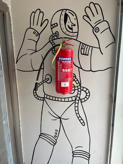 A red "FIRE KILLER" powder fire extinguisher is mounted on a white wall in Isasi, Nigeria. The extinguisher is centrally positioned over a black line-art mural depicting a stylized astronaut or spaceman figure with raised arms and a detailed helmet. A yellow strap secures the extinguisher to the wall. Its black hose extends from the top, curving downward, appearing to connect to the drawn figure's neck area. Metal door or frame elements are visible vertically along the left and right edges of the white wall section.