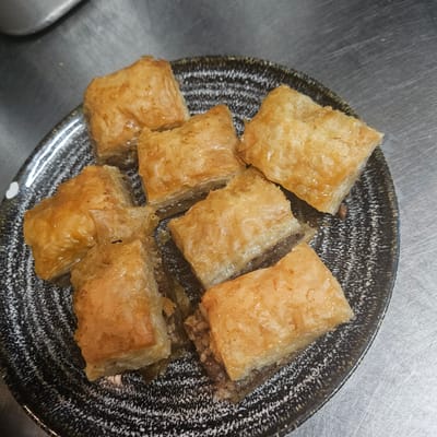 A plate of baklava sits on a metallic surface. The plate is round, with a dark grey rim and a speckled white and grey pattern on the inside. Seven pieces of golden-brown baklava are arranged on the plate. The environment is a kitchen or food preparation area, evidenced by the metallic surface. The location, Fehmarn, Germany, is external information, not visually evident in the image. There are no people in the image.