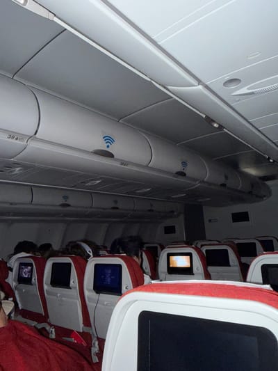 An interior view of an airplane cabin, likely an economy class section, is captured with a dim, ambient lighting, suggesting a night flight or relaxed cabin atmosphere. The image is oriented vertically, showing the length of the cabin from a perspective near the window or aisle, looking towards the opposite side.
On the right side of the frame, several rows of aircraft seats are visible. The seats feature red fabric upholstery and white or light-colored frames, each equipped with a personal entertainment screen on the seatback. Some screens display content, including one with illuminated text or an image, while others are dark or off. Passengers are seated, with their heads and shoulders partially visible, appearing to be resting or engaged with the in-flight entertainment.
The left side of the frame showcases the overhead bins and ceiling panels of the aircraft. These are predominantly white or light grey. Multiple blue Wi-Fi symbols, depicting three curved lines radiating upwards, are affixed to the underside of the overhead bins, indicating the availability of in-flight internet connectivity. One overhead bin clearly shows the seat number "39 A". The overall impression is a typical commercial flight cabin during a period of low activity, with passengers settled in their seats. The provided location context, Mysy, Russia, is not directly discernible from the interior of the aircraft.