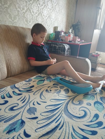 A young boy sits on a couch in a room with patterned wallpaper. He is wearing a dark blue polo shirt with red trim and light-colored shorts. The boy is looking down, possibly at something on his lap or on the floor in front of him. His legs are extended, and his feet are bare.

In front of the boy, on a patterned blanket covering the couch and a table, is a blue electronic device with a black grip. The blanket has a floral pattern in shades of blue and white.

In the background, a desk is visible with various items on it, including cups, bottles, and what appears to be a stack of boxes. A chair or a cabinet with drawers is also present. The lighting suggests it is daytime. The room appears to be a living space or a bedroom in a home in Kingisepp, Russia. No specific emotion is clearly discernible on the boy's face, but his posture suggests a relaxed or focused state. There is no visible text in the image.