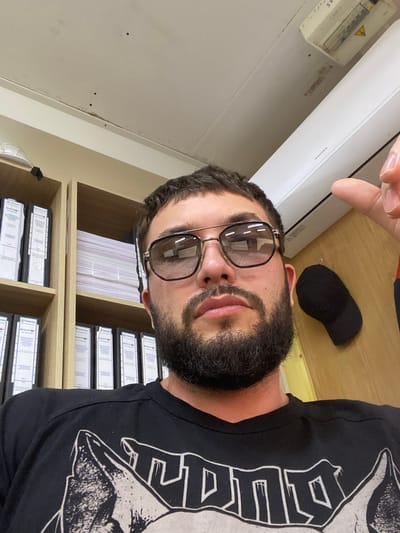 A man with a beard and sunglasses is taking a selfie in what appears to be an office. He is wearing a black t-shirt with a graphic design. Behind him, there are shelves filled with binders and papers. An air conditioner unit is mounted on the wall above the shelves. A black baseball cap is hanging on the wall to the right of the man. The image does not provide enough information to determine the specific location within Luzhitsy, Russia.