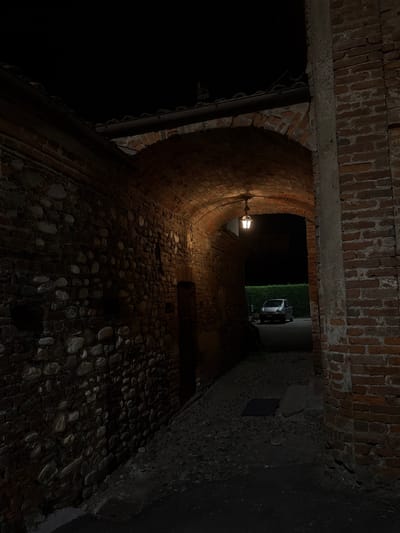This is a nighttime photograph of a narrow stone alleyway in Lagnasco, Italy, possibly a historic district or a courtyard. The alleyway is covered by a brick archway, under which a single lantern hangs, casting a warm light. The walls of the alley are constructed of rough-hewn stones and bricks, with a cobblestone pavement. In the distance, beyond the archway, a silver compact car is parked in front of a neatly trimmed hedge. The scene is dimly lit, with shadows adding to the atmosphere of the quiet night.