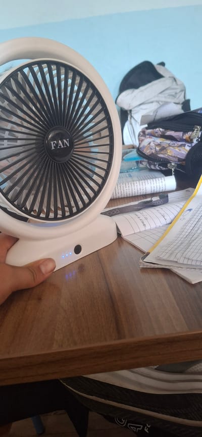 A close-up, eye-level shot captures a white portable fan placed on a wooden desk. The fan, a prominent object, features a black grill with the word "FAN" in white lettering at its center. To the right of the fan, a stack of open notebooks and papers are scattered across the desk, with a yellow ruler lying on top of one notebook. Further in the background, a partially visible backpack and other items are present. The desk surface appears to be made of polished wood with a visible grain. The lighting suggests an indoor setting, possibly in a home or office. The background wall is a light blue. The bottom left corner of the image shows a hand, with fingers extended and resting on the desk near the fan's base, which has four blue indicator lights.
