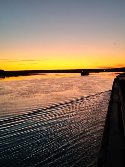 The image depicts a broad river at sunset, with a vessel on the horizon and a dark structure on the right bank, likely a quay or a large boat. The sky is a gradient of orange, yellow, and blue, with a thin strip of land visible in the distance. The water reflects the colors of the sky, creating shimmering patterns. The perspective suggests the photo was taken from a moving vessel, as evidenced by the wake behind it. The scene is tranquil and atmospheric, characteristic of a sunset over a waterway.

While the image captures a river scene at sunset, it is impossible to definitively identify it as being in Yaroslavl, Russia, or any specific city without further contextual information. The presence of a river, a quay or boat, and a sunset are common worldwide. Therefore, a precise location in Yaroslavl cannot be determined solely from the visual content of the photograph.