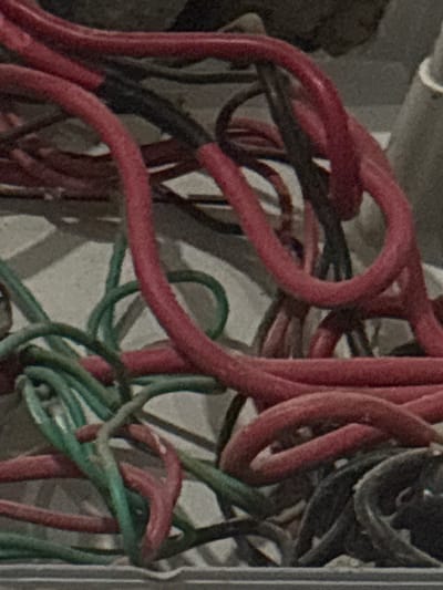 The image displays a close-up, static shot of a dense tangle of electrical wires. The primary subjects are numerous red-sheathed wires, heavily intertwined with several green and dark/black wires. The wires appear to be of various gauges and are coiled and looped irregularly, suggesting a chaotic or exposed wiring installation rather than a neatly organized system.

The background consists of muted gray and off-white tones, possibly indicating a wall or the inside of an electrical box or conduit, with subtle textures and shadows adding to the depth of the scene. No specific activity is taking place in the image; it is a snapshot of the wires as they are.

Notable details include the clear distinction between the red, green, and dark wire colors. The image has a visible graininess or noise, hinting at potentially low-light conditions or a camera with limited resolution. There are no people, explicit emotions, time of day indicators, weather conditions, or visible text within the frame. The location context of Angwan Sayawa, Nigeria, is not visually discernible from the image itself.