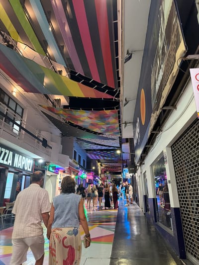 The image depicts a vibrant, narrow pedestrian street lined with shops and decorated with colorful banners. Several people are seen walking along the street, appearing to be tourists or shoppers. The banners, predominantly in rainbow stripes and geometric patterns, create a festive and lively atmosphere. Above the banners, architectural details of the buildings are visible, including metal grating on the ceiling and exposed structural elements. Some shops have illuminated signs, suggesting the scene takes place in the evening or at night, or perhaps in an indoor market. The flooring is a dark, reflective surface, possibly asphalt, with colorful painted patterns. Signs such as "PIZZA NAPOLI" are visible, indicating a commercial area with food establishments. The overall impression is one of a bustling, colorful, and inviting urban environment, likely a popular tourist destination given the context of Sant Antoni De Portmany, Spain. The people appear relaxed and are engaged in leisurely activity.