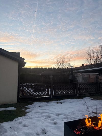 The image depicts an outdoor scene in Yanino-1, Russia, under a twilight sky with scattered altocumulus clouds exhibiting hues of blue, pink, and orange near the horizon. A prominent contrail extends vertically through the upper center of the sky.

In the immediate foreground, to the bottom right, a dark, square metal fire pit contains visible flames and embers. The ground is largely covered with an irregular layer of melting snow, interspersed with patches of green grass, particularly on the left. Several small, amorphous snow piles are scattered across the white surface.

Towards the midground, a dark brown wooden fence with a lattice pattern runs horizontally. A dark lamp post with a lantern-style top is positioned just behind this fence on the left side of the frame. Beyond the fence, further structures are visible. On the far left, a portion of a light-colored building with a dark roof and gutter system is present. To the right of the lamp post, another structure features a dark roof and a tall, dark chimney-like vent. A tarp-covered object or structure is situated to the right of this chimney.

The background consists of a dense line of bare deciduous trees and some evergreen foliage, silhouetted against the bright horizon. Additional building rooftops are partially visible among the trees. No individuals are discernible within the frame.