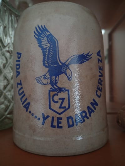 The image shows a close-up of a beige ceramic stein with a blue emblem of a flying eagle and text. The text, arranged in a circular pattern, reads "PIDA ZULIA... Y LE DARAN CERVEZA". This translates to "Ask for Zulia... and they will give you beer". The stein is likely a souvenir or promotional item related to Cerveza Zulia, a beer brand from Venezuela. The background is blurred, but suggests an indoor setting, possibly a shelf or display area. The lighting is natural, coming from the right, casting a subtle sheen on the stein's surface, highlighting some texture and minor imperfections in the ceramic. The overall impression is of a still life, focusing on the details of the stein and its inscription. There is no indication of the city of Valencia, Venezuela, in the image itself; this context is external to the visual information provided.