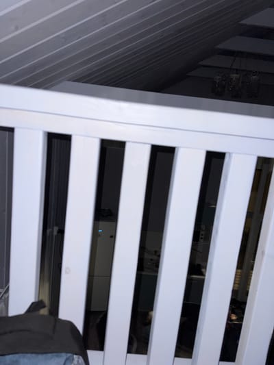 The image captures a view through white wooden slats, likely part of a crib or railing. Beyond the slats, a dimly lit interior is visible. In the background, a ceiling with wooden paneling slopes upwards, and a chandelier with multiple candle-like lights hangs from it. To the left, a light-colored vertical appliance, possibly a dehumidifier or air purifier, is present, with a blue indicator light on its front. Further in the background, other indistinct objects and shadows suggest a cluttered or storage-like space. The overall scene conveys a sense of being indoors, with limited light and a focus on the wooden structure in the foreground. The location is Doma 1096 Km, Russia.