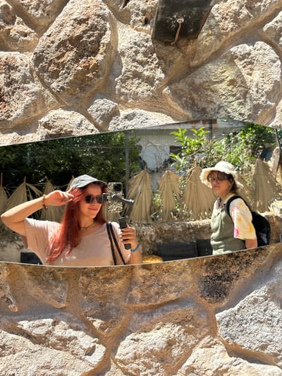 The image captures a vertically oriented mirror, reflecting two women and a vibrant outdoor scene, all framed by a rugged, light-colored stone wall. The location is Kaş, Turkey.

In the foreground of the mirror's reflection, a woman with long, reddish-brown hair, wearing sunglasses and a dark cap, smiles at the camera. She is clad in a light-colored t-shirt and holds a smartphone mounted on a gimbal in her right hand, suggesting she is taking a selfie or recording a video. Her left hand is raised, making a 'peace' sign or similar gesture. Further back in the reflection, partially obscured, another woman is visible, wearing a light-colored, fluffy hat, glasses, and a light-colored top over a green jacket.

The background reflected in the mirror consists of lush green foliage, possibly trees and shrubs, interspersed with what appear to be dried palm fronds or large, fan-shaped leaves, hinting at a Mediterranean or arid garden environment.

The mirror itself is bordered on both sides by a textured stone wall, composed of irregularly shaped, light-colored rocks, creating a rustic and natural aesthetic. The strong sunlight casts clear shadows on the stone, indicating a bright, sunny day. The overall mood conveyed by the smiling woman is relaxed and cheerful. No discernible text is visible in the image.