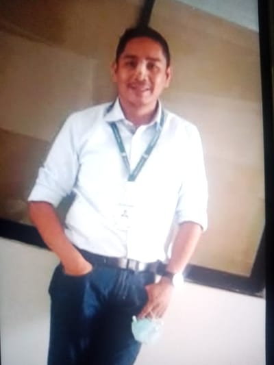 A man stands indoors. He is wearing a light blue, collared shirt with the sleeves rolled up, dark blue jeans, a black belt, and a wristwatch on his left wrist. He has a lanyard around his neck. His left hand is in his left pocket, and he is holding a light blue face mask in his right hand. The background appears to be a wall with a dark, horizontal stripe. The location is Santa Cruz de la Sierra, Bolivia.