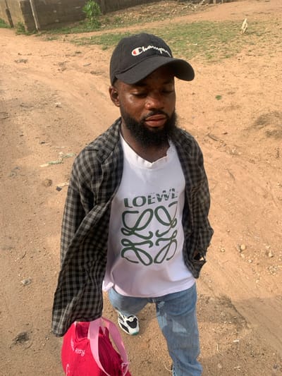 A male individual is depicted standing on a reddish-brown dirt path in Eika-Adagu New Layout, Nigeria. He is wearing a black baseball cap with "Champion" embroidered in white and red, a white t-shirt featuring a green "LOEWE" text logo above a stylized green interlocking "L" pattern, an open black and white checkered long-sleeve shirt, blue jeans, and black and white athletic shoes with light blue accents. His facial hair includes a beard and mustache. He holds a bright pink backpack or small bag, partially visible with a "Hello Kitty" logo, in his left hand. His eyes appear closed or directed downwards, with his head slightly tilted. The ground is unpaved, consisting of packed dirt with small stones and sparse green vegetation. In the background, a structure of concrete blocks is visible on the left, and further right, a faint white bird (possibly a chicken) is present amidst the vegetation and bare ground. The scene is illuminated by natural daylight.