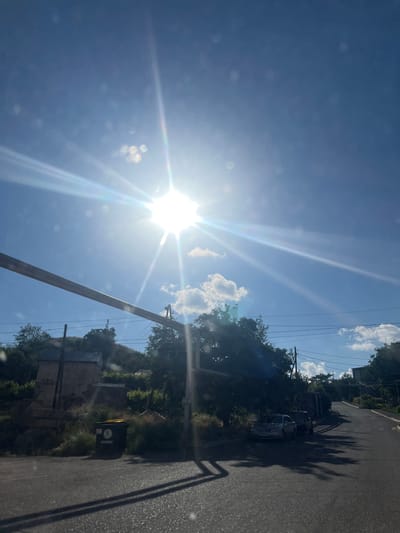 The image depicts a sunny day in Gladzor, Armenia. The scene is a street with a clear blue sky and bright sunlight, creating lens flare and god rays. There are trees and some houses visible along the side of the road. A few cars are parked on the side of the street, and a trash bin is also present. A utility pole with wires runs along the side of the road, extending into the frame. The overall impression is a typical daytime scene in a semi-rural or residential area. The time of day is clearly during daylight hours, likely mid-morning to afternoon, given the intensity of the sun and the shadows. The weather appears to be clear and sunny, with a few scattered clouds. There are no people visible in the image. No text is discernable in the image.