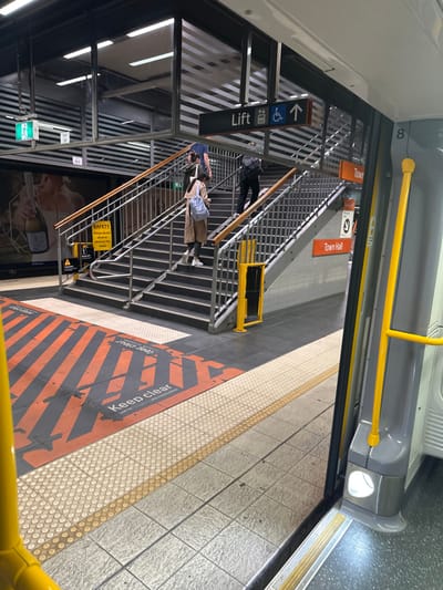 A sideways view from inside a train at a station, focusing on stairs and signs. The scene features several people on the stairs, with a lift sign pointing upward. Signs for "Town Hall" are visible. Safety warnings and "Keep Clear" markings are painted on the platform. The train's yellow handrail and number "8" are also visible. An advertisement is positioned near the bottom of the stairs.