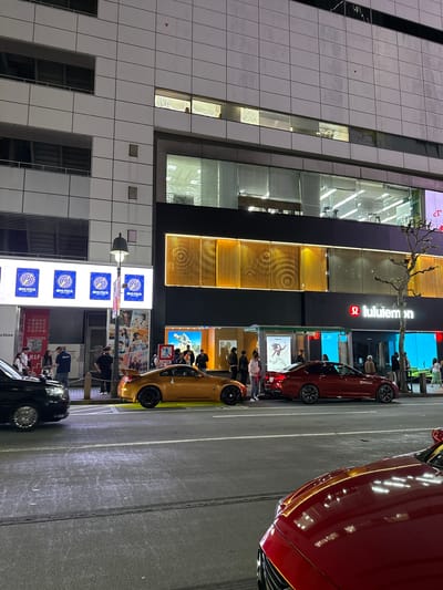The image depicts an urban street scene at night, likely in Shibuya, Japan. Visible are: a multi-story building with the "lululemon" logo, several parked cars (one red, one gold, and one black), and advertisements. Pedestrians are seen near the storefront, engaging in activities such as looking at merchandise and standing nearby. A series of vertical advertisements featuring the "ONE PIECE" franchise are also displayed. The lighting is a mix of building lights, advertisement lights, and car headlights.