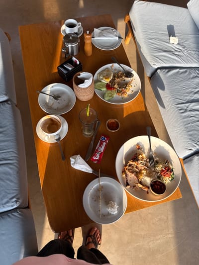 The image shows a table with leftover food and drinks, suggesting a meal has recently concluded. On the wooden table are two plates with rice, meat, and vegetables, as well as an empty coffee cup and a glass with a lime wedge. A silver teapot, salt and pepper shakers, and a small basket with sugar packets are also present. The scene appears to be an outdoor dining area, possibly a cafe or restaurant in Banjar, Indonesia, as indicated by the presence of light-colored outdoor seating and the warm, natural lighting. The overall impression is one of a relaxed, post-meal setting.