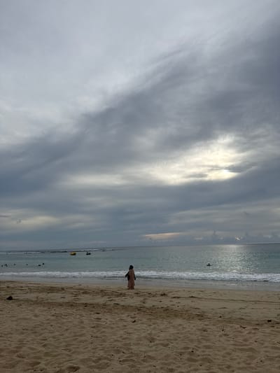The image shows a beach scene in Meunasahbale, Indonesia, under a cloudy sky. The foreground is a sandy beach with tire tracks visible. In the midground, the ocean meets the shore with gentle waves. A single person, wearing a long, light-colored garment, stands facing the water. Further out in the ocean, several people are visible. Some are on a yellow banana boat, and others appear to be on jet skis or engaged in water activities. The sky is filled with dramatic, grey clouds, with a bright patch of sunlight breaking through in the upper right portion of the image, casting a reflection on the water. The overall atmosphere is one of an overcast day at the beach with moderate activity.