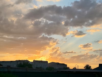 This media file captures a scenic view of apartment buildings against a dramatic sunset sky. The scene is set in Taldykorgan, Kazakhstan. The time of day is clearly dusk, with the sun low on the horizon, casting a warm, golden-orange glow across the clouds and illuminating the buildings. The sky is a canvas of varying cloud formations, from dense, grey masses to wispy, illuminated edges, with rays of sunlight piercing through them, creating a sense of depth and wonder.

The foreground features the rooftops of low-rise structures, possibly sheds or garages, and a dark, green fence, suggesting a residential or suburban area. Behind them, several multi-story apartment buildings, typical of Soviet-era architecture, are silhouetted against the vibrant sky. The buildings are grey and feature numerous windows, some of which may reflect the sunset's colors. A few trees are interspersed between the buildings and behind the lower structures, their branches dark against the bright sky. Streetlights and utility wires are also visible, adding to the urban setting.

The overall mood is peaceful and serene, evoking the beauty and calm of a day ending. The vibrant colors of the sunset, transitioning from fiery orange to softer yellows and blues, create a visually striking contrast with the muted tones of the buildings and foreground. There is no visible text in the image.