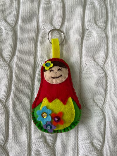 A handmade felt keychain resembling a Russian doll lies on a white cable-knit sweater. The doll has a red body, a cream-colored face with embroidered features, a yellow loop for the keychain ring, and colorful felt flower embellishments. It is photographed indoors. No location is recognizable in this scene. Лесной Пейзаж Кп (шарапово, Russia is not visible.