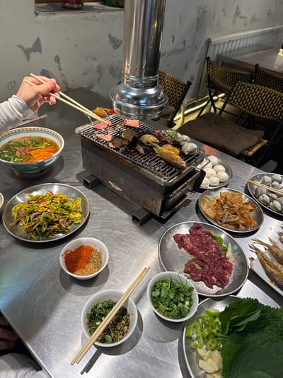 The image depicts an overhead view of a Korean barbecue-style meal taking place in a restaurant located in Lishan District, China. A silver rectangular tabletop grill, centered in the frame, is actively cooking various items over coals, including cuts of beef, whole small fish, shrimp, mushrooms, and clams. A large, retractable metal exhaust hood extends downwards over the grill.

On the left, a person's left hand is visible, holding a pair of light-colored chopsticks to manipulate meat on the grill. Adjacent to the hand is a colorful bowl containing a noodle or soup dish garnished with green onions and shredded carrots.

The meal is spread across a metallic, presumably stainless steel, table. Other dishes present include:
*   A silver plate of stir-fried green peppers mixed with meat or vegetables.
*   Two small white bowls containing dry spices: one with red chili flakes and what appears to be ground sesame, and another with an unidentified dry mix.
*   A small white bowl containing a liquid dipping sauce with chopped green onions and a pair of chopsticks resting across it.
*   A small white bowl filled with fresh chopped cilantro and green onions.
*   A silver plate piled with raw, thinly sliced red beef.
*   A silver plate containing raw, marinated squid or octopus pieces.
*   A silver plate with multiple raw clams in their shells.
*   A white rectangular plate holding several small, whole raw fish.
*   A silver plate with whole white button mushrooms.
*   A silver plate containing large green leaves (possibly lettuce or perilla), sliced green bell peppers, and peeled garlic cloves.

In the background, two dark woven chairs with beige seat cushions are visible, and the restaurant's wall features a distressed, light-colored finish. The scene suggests a communal dining experience where patrons cook their own food at the table.